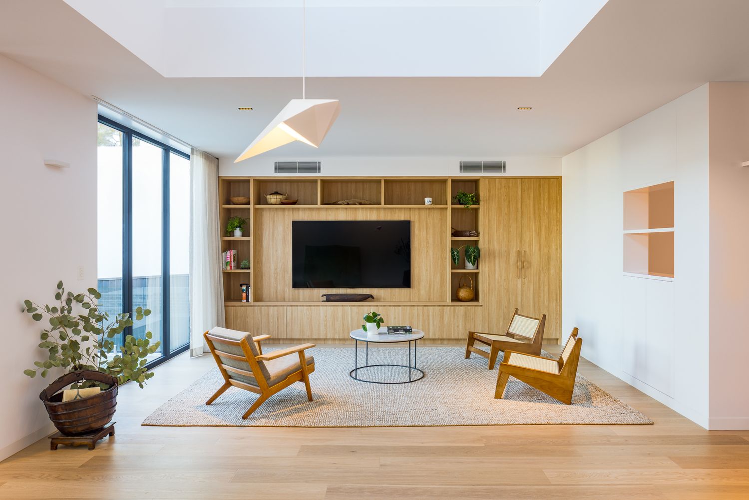 Bright open living room with timber wall unit, built-in television, mid-century armchairs, round coffee table and floor-to-ceiling windows.