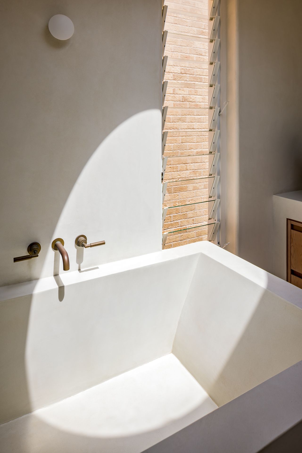 Close-up of a modern plaster bathtub with brass taps, louvre window with brick view and sunlight patterns on the wall.