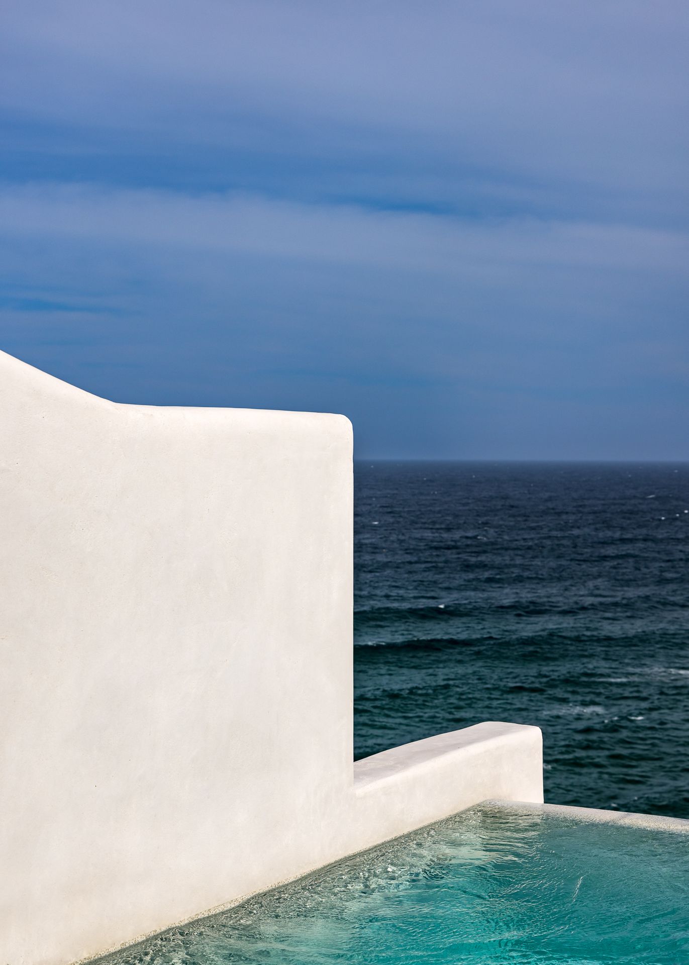 White Mediterranean-style wall beside a turquoise infinity pool overlooking the deep blue ocean under a cloudy sky.