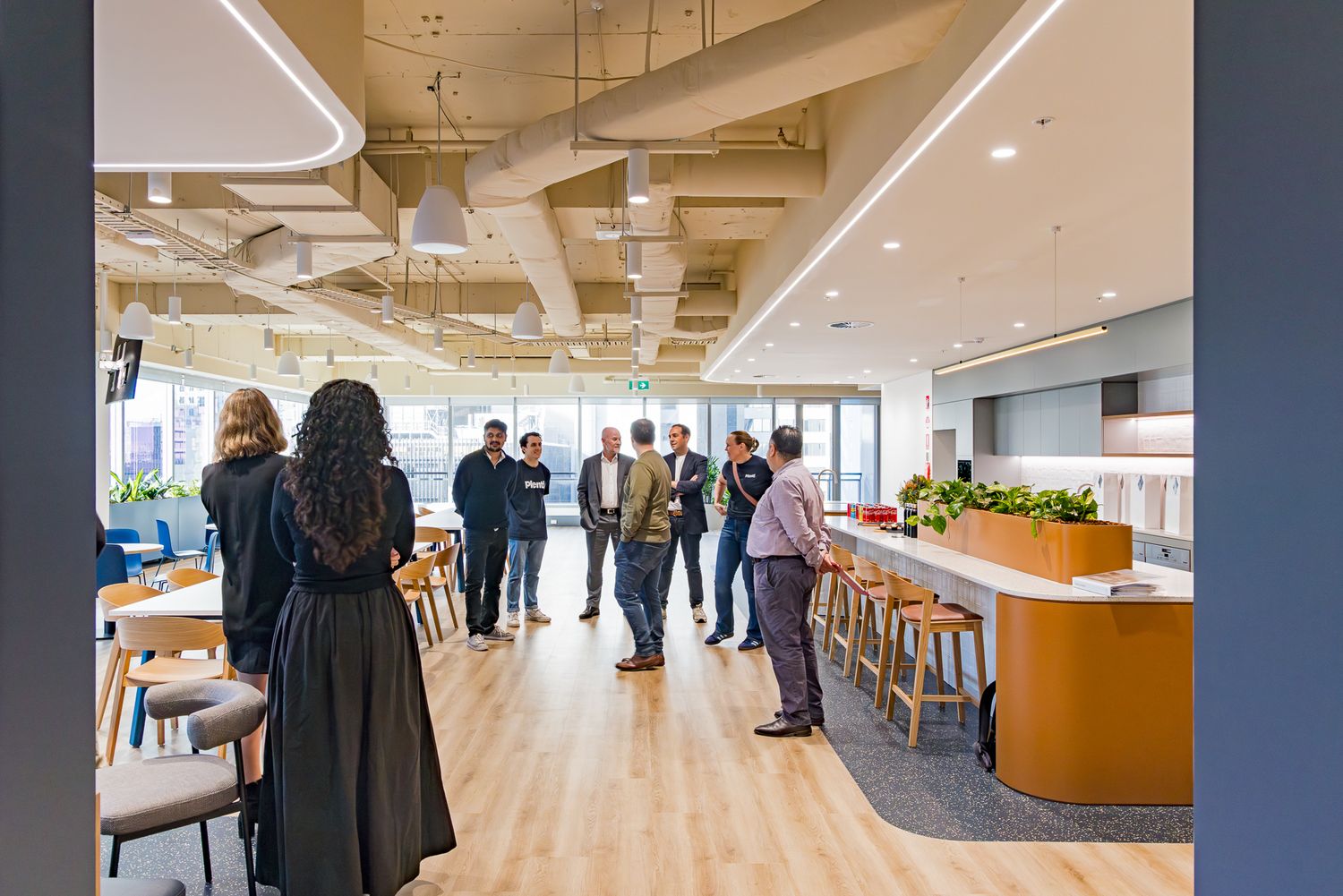 Group of employees chat in an open office kitchen and break area with exposed ceilings, wood floors and a wooden counter with stools.