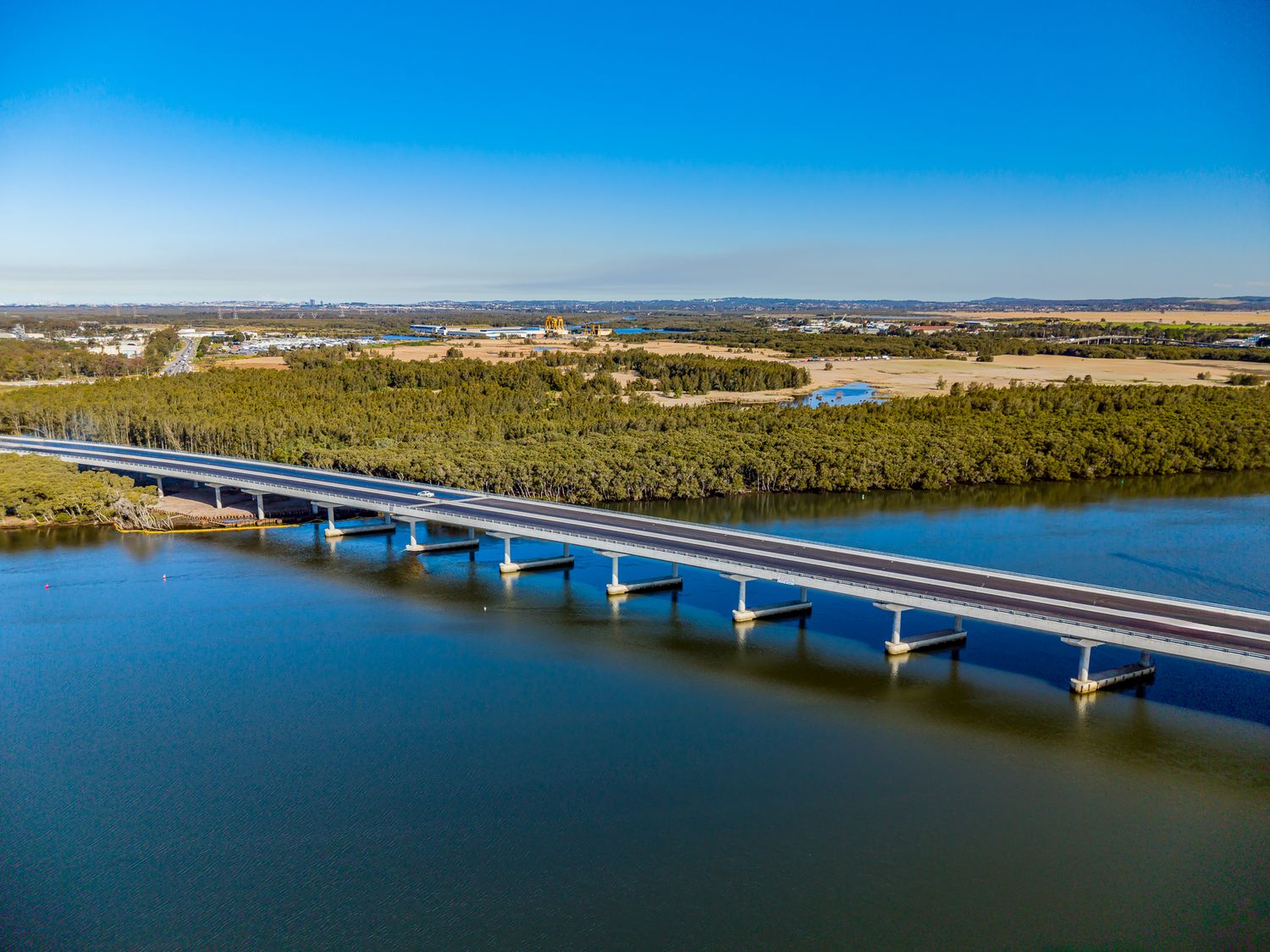 Aerial view of a long concrete highway bridge crossing a calm blue river lined with green bushland under a clear sky.