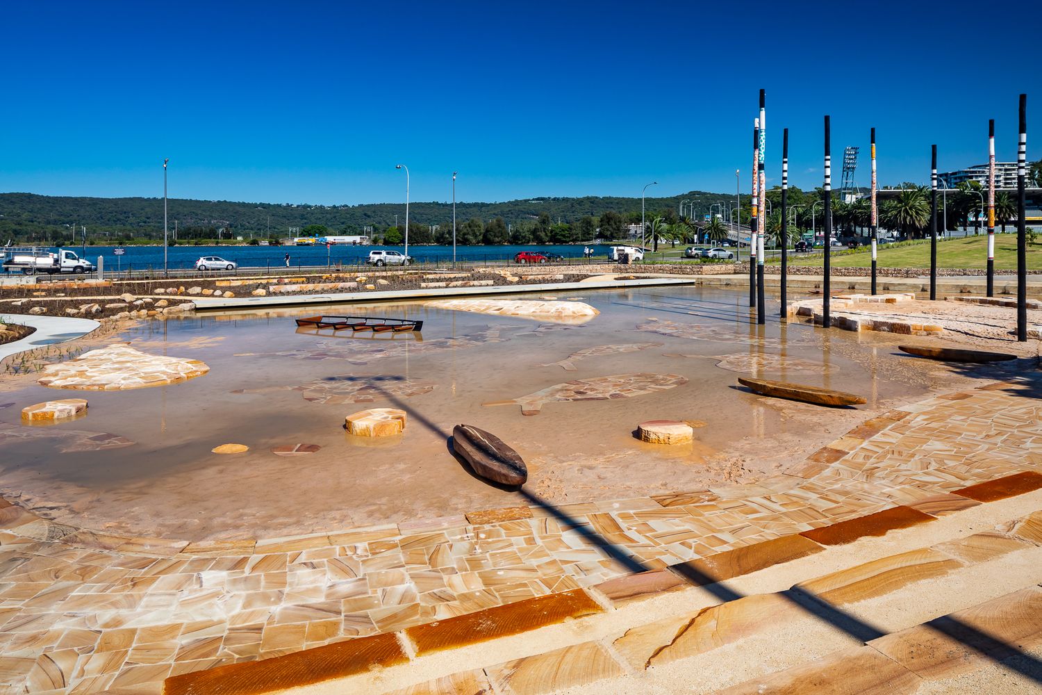 A wide shallow water play area with sandstone paving and colorful poles overlooks a calm waterfront and distant hills.