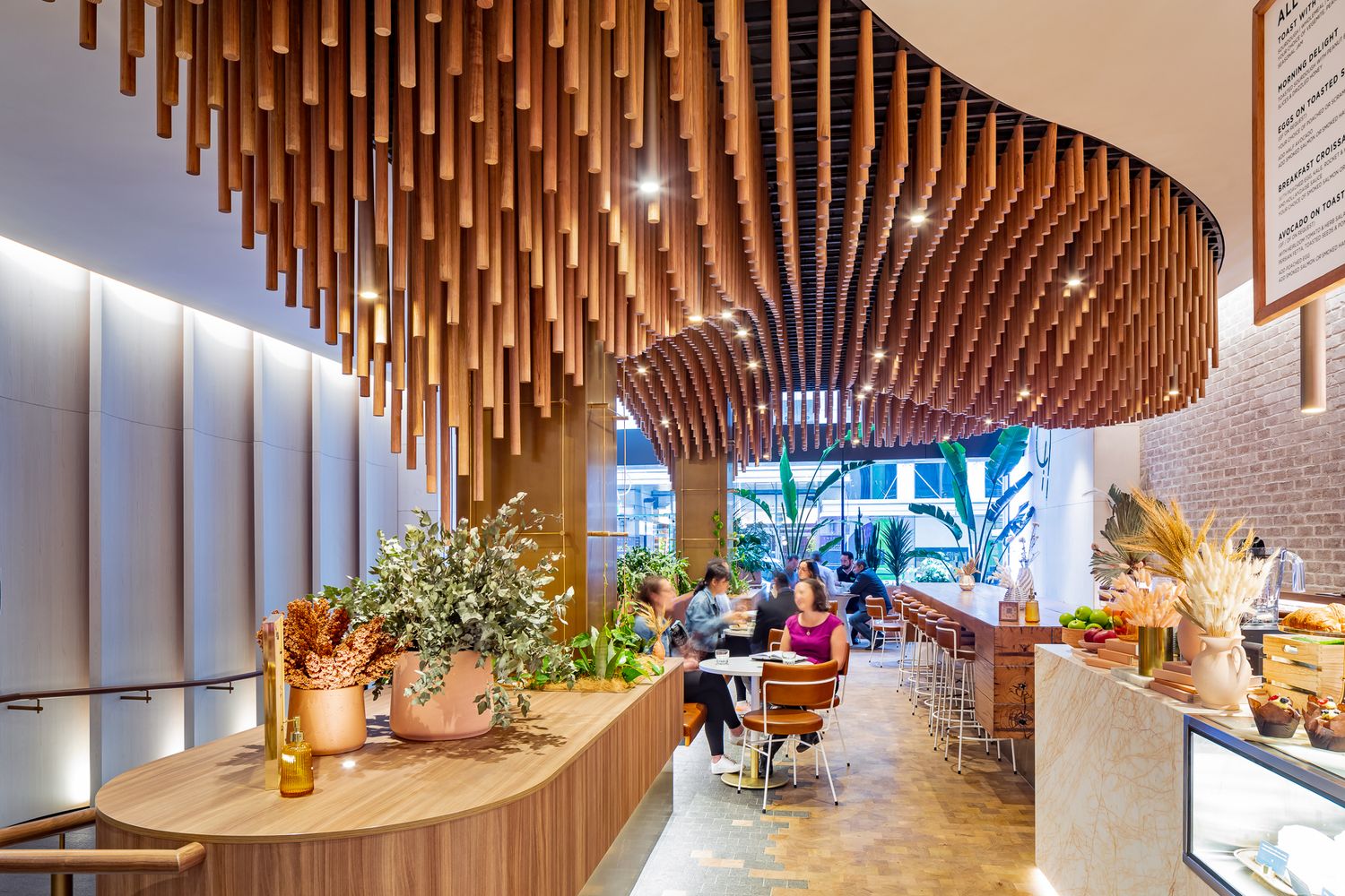 Modern cafe interior with a sculptural wooden ceiling of hanging timber battens above a curved counter, potted plants and diners.