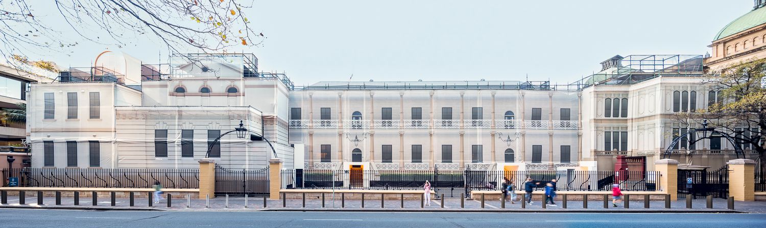 Wide view of a long historic building facade with arched entrance and iron fence, with pedestrians walking past on the street.