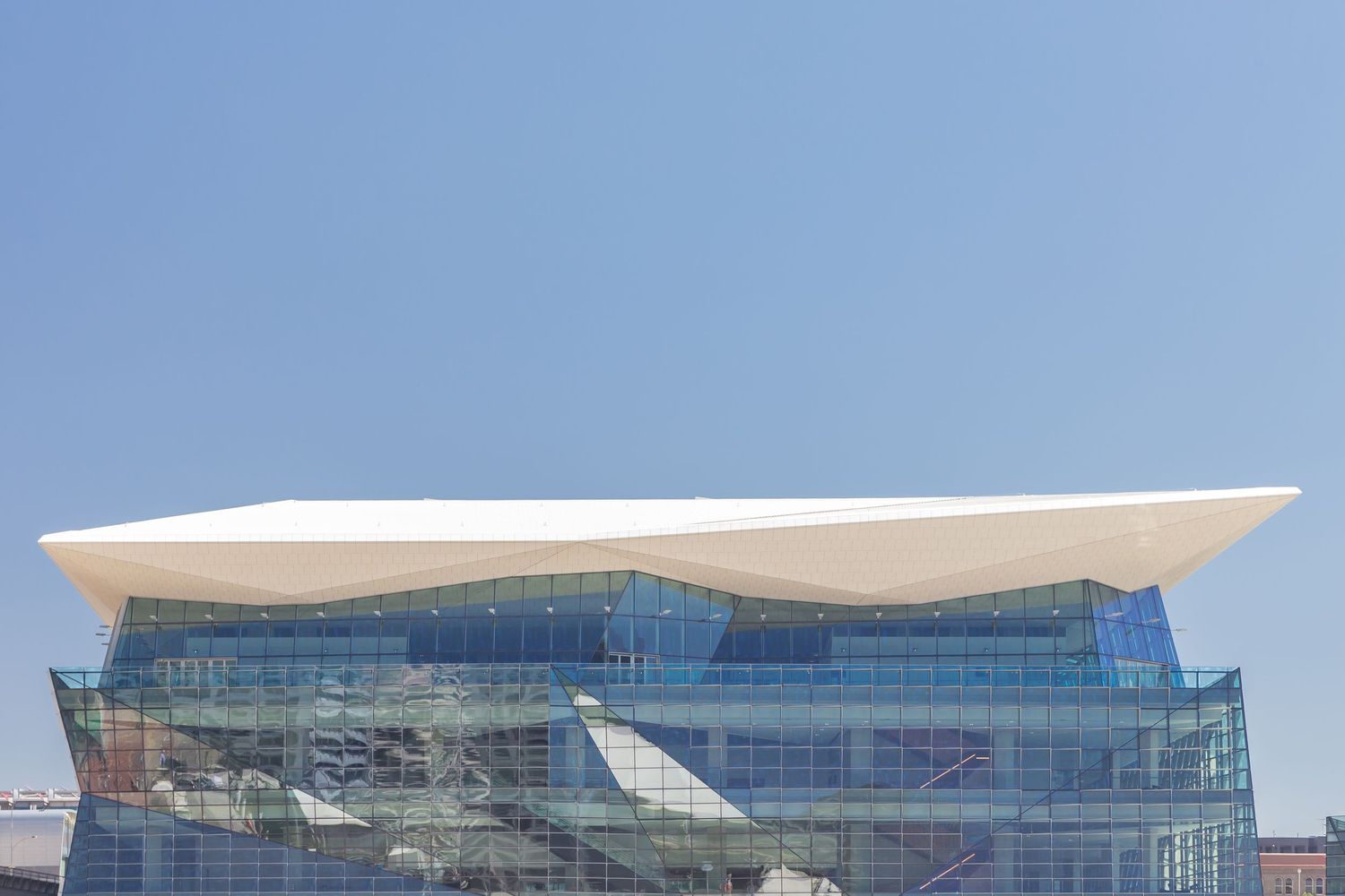 Contemporary glass building topped with a sweeping white angular roof against a clear blue sky.