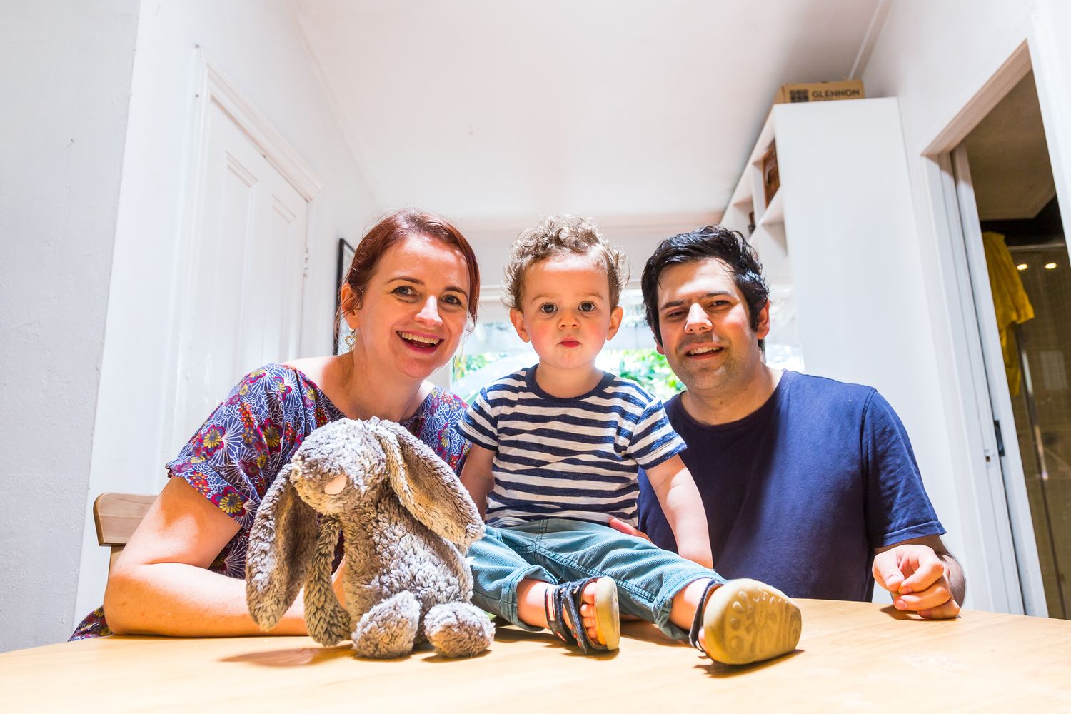 Smiling mother, father and toddler son sit together at a wooden table at home with a plush grey bunny toy in the foreground.