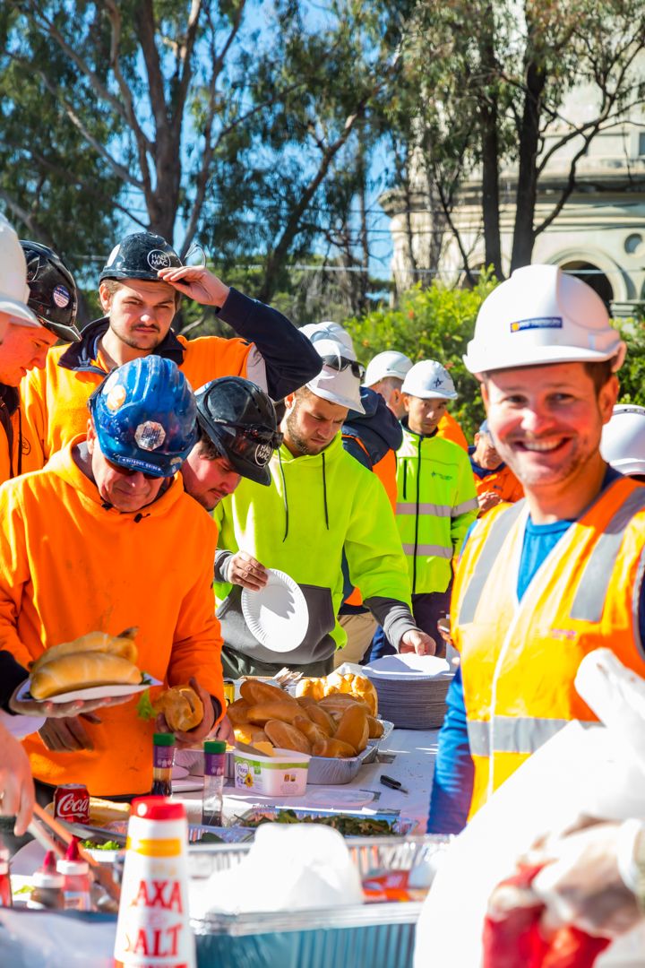 Smiling construction workers in hi-vis gear and hard hats gather around a outdoor catering table serving burgers and food on a sunny day.