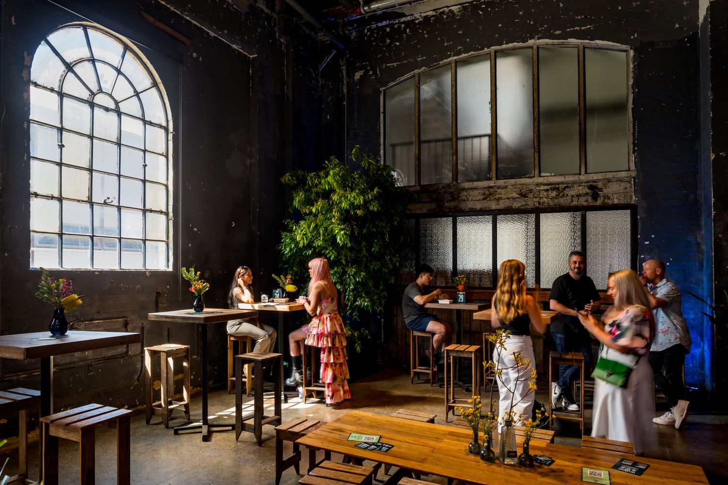 Patrons chat at tall tables and stools in a rustic industrial venue with exposed brick walls, large arched windows and a leafy plant.
