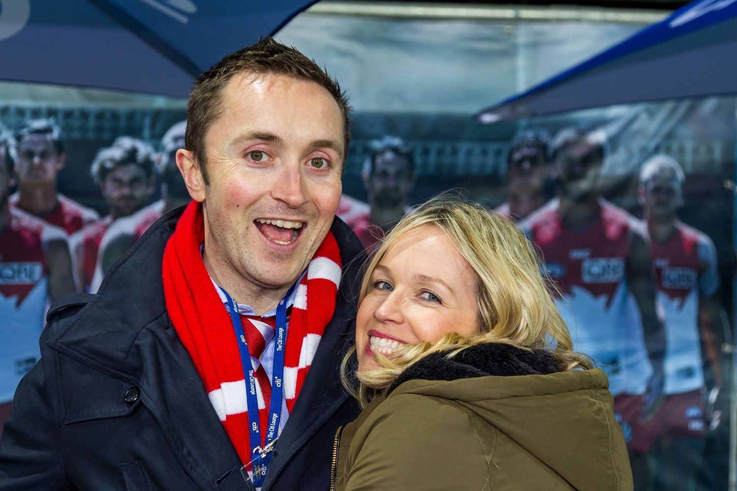 A smiling man and woman pose together at a stadium, with the man wearing a red and white scarf and lanyard.