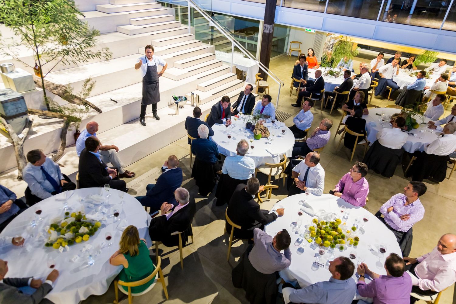 Guests sit at round tables with floral centerpieces while a chef in an apron stands on steps at a lunch event.
