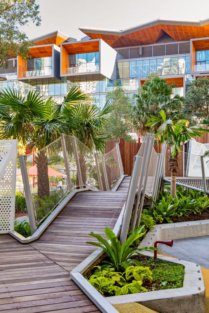 Timber boardwalk and landscaped garden with palm trees leading to a modern apartment building with orange timber-clad rooflines.