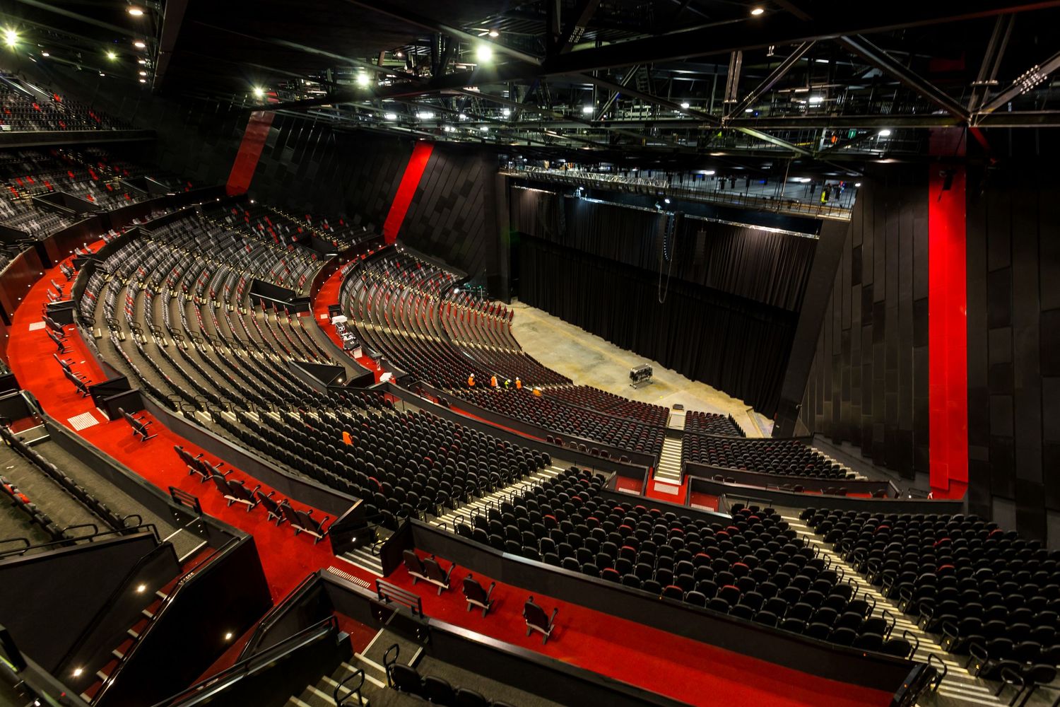 Interior of a modern arena with tiered black seating, red accent stairs and walls, and a central event floor under steel roof trusses.