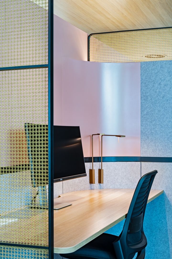 Private office nook with a wood desk, black monitor, brass desk lamps and chair, framed by perforated metal screens and pink felt panels.