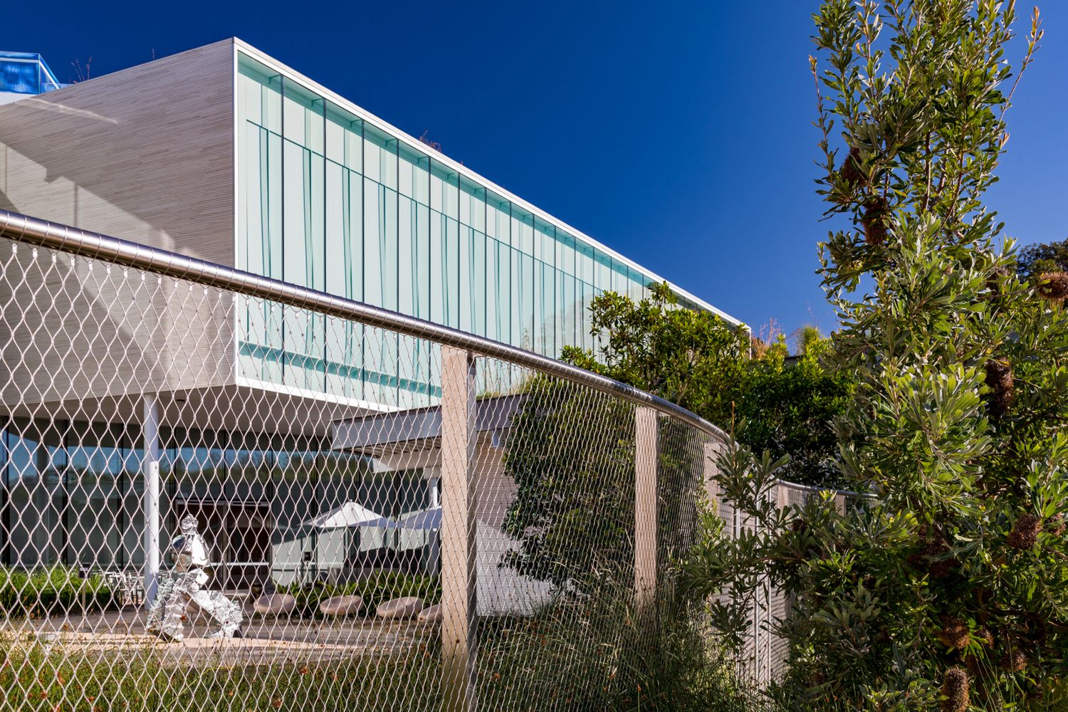 Modern building with a glass and metal facade seen through steel mesh fencing and green trees under a bright blue sky.