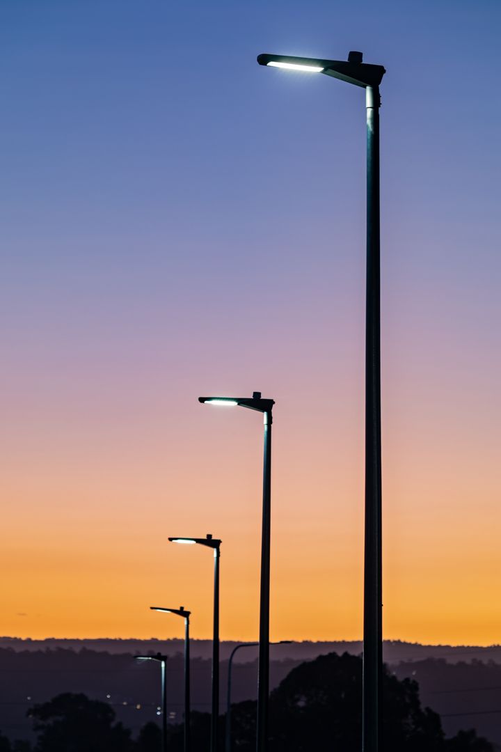 Row of illuminated street lamps in silhouette against a gradient dusk sky fading from deep blue to orange over distant tree-covered hills.