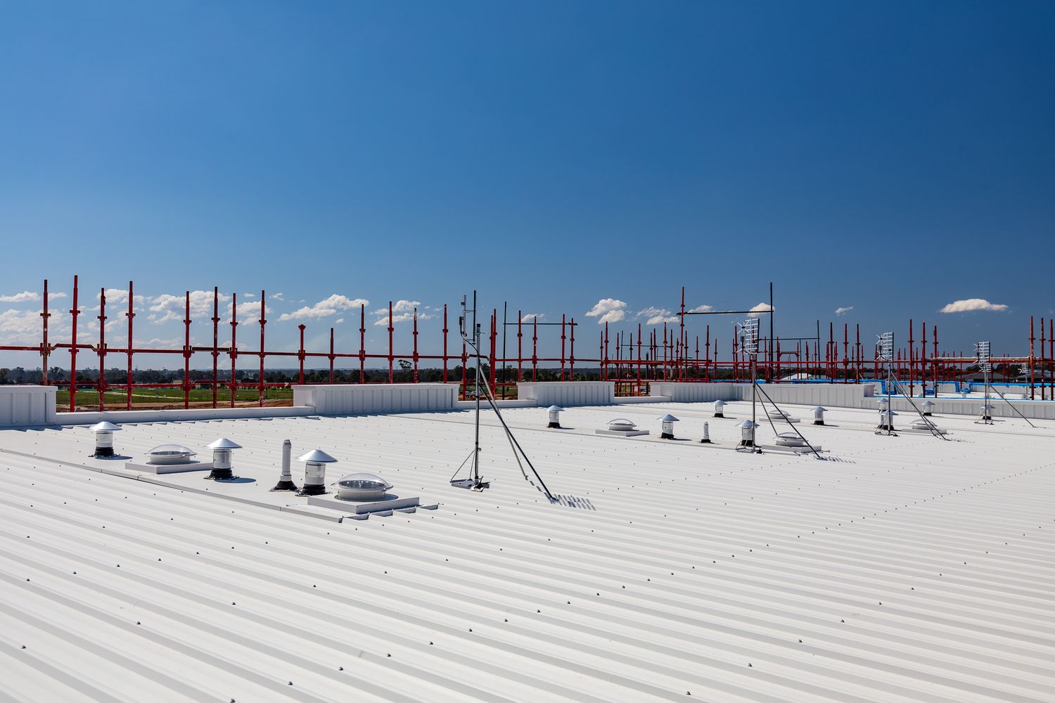 Industrial building with a white corrugated metal roof, rooftop vents and red steel framework against a clear blue sky.