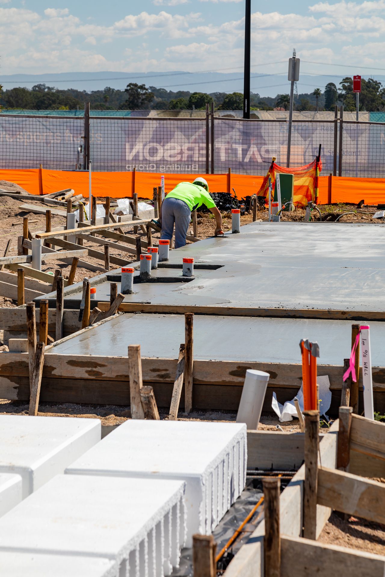 Construction worker in hi-vis gear setting up concrete slab formwork with timber stakes beside stacked white polystyrene waffle pods.