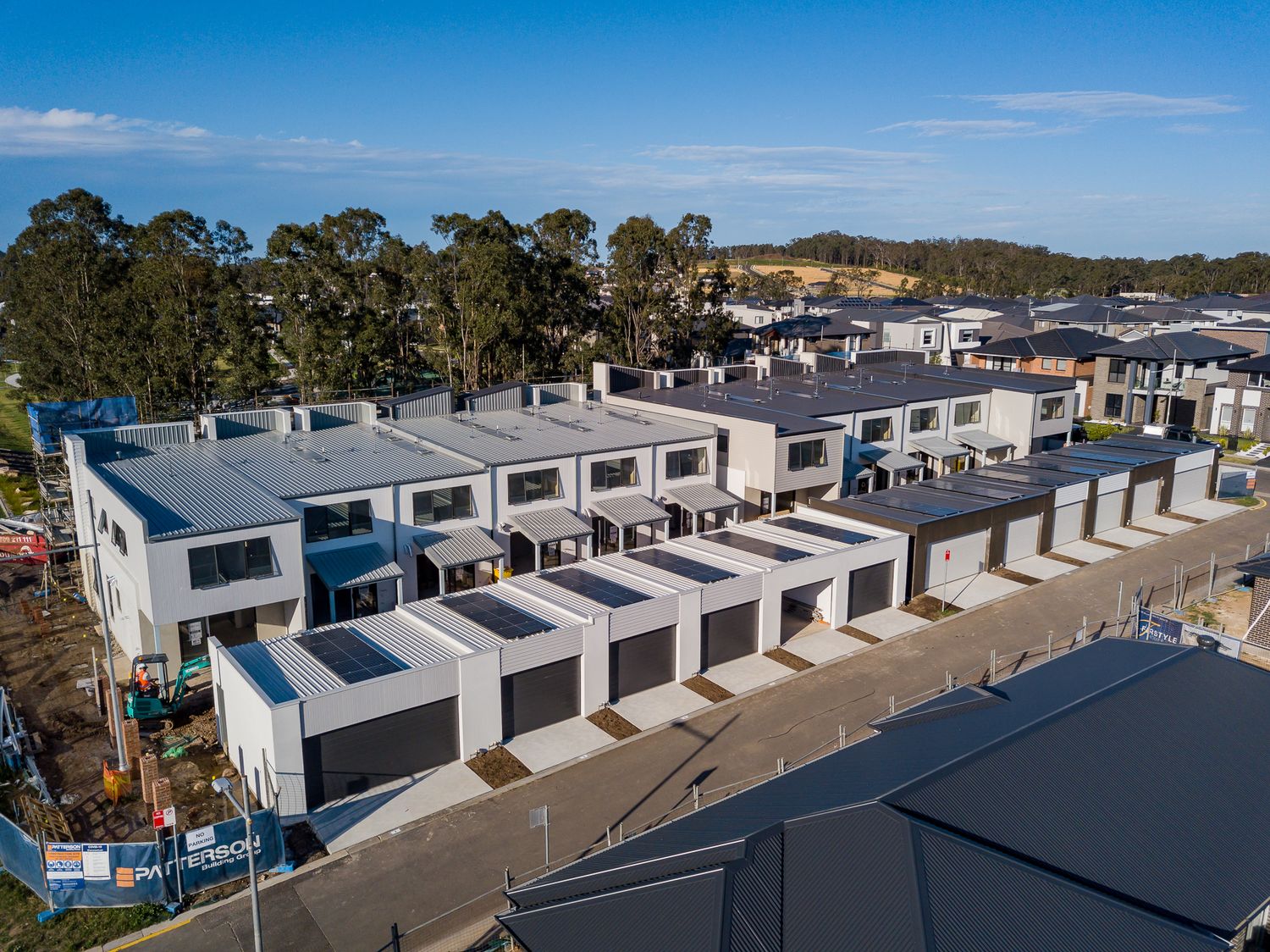 Aerial view of a new townhouse development with rows of modern homes, garages, and trees on a hillside in the background.