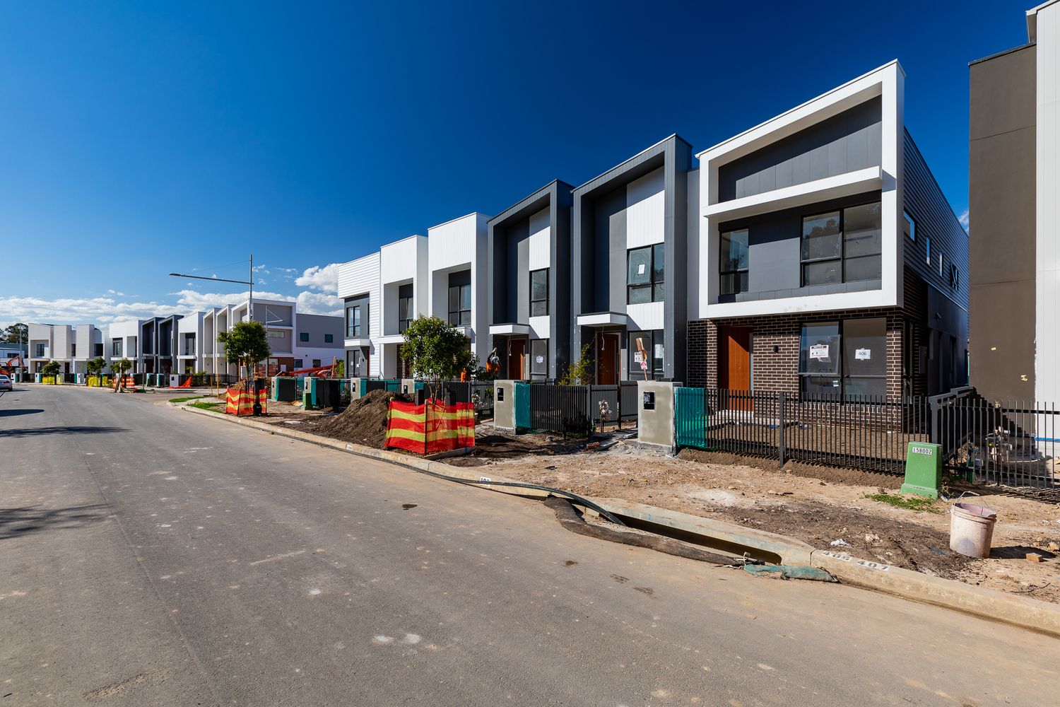 Street view of newly built modern townhouses with a young tree and construction fencing beneath a clear blue sky.