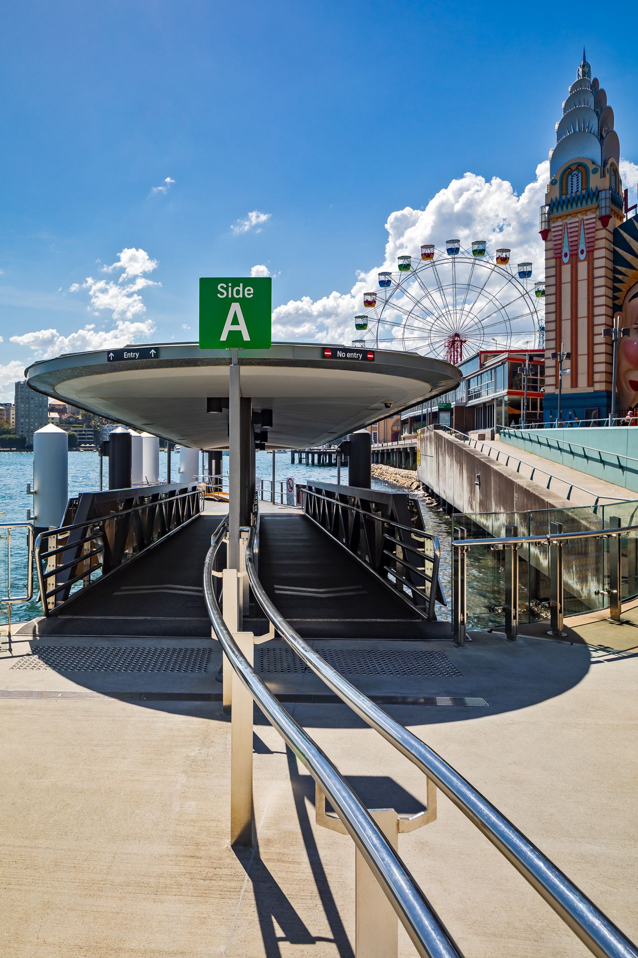 Ferry wharf entrance marked Side A with a curved canopy, overlooking Luna Park's Ferris wheel and ornate tower on a sunny day.