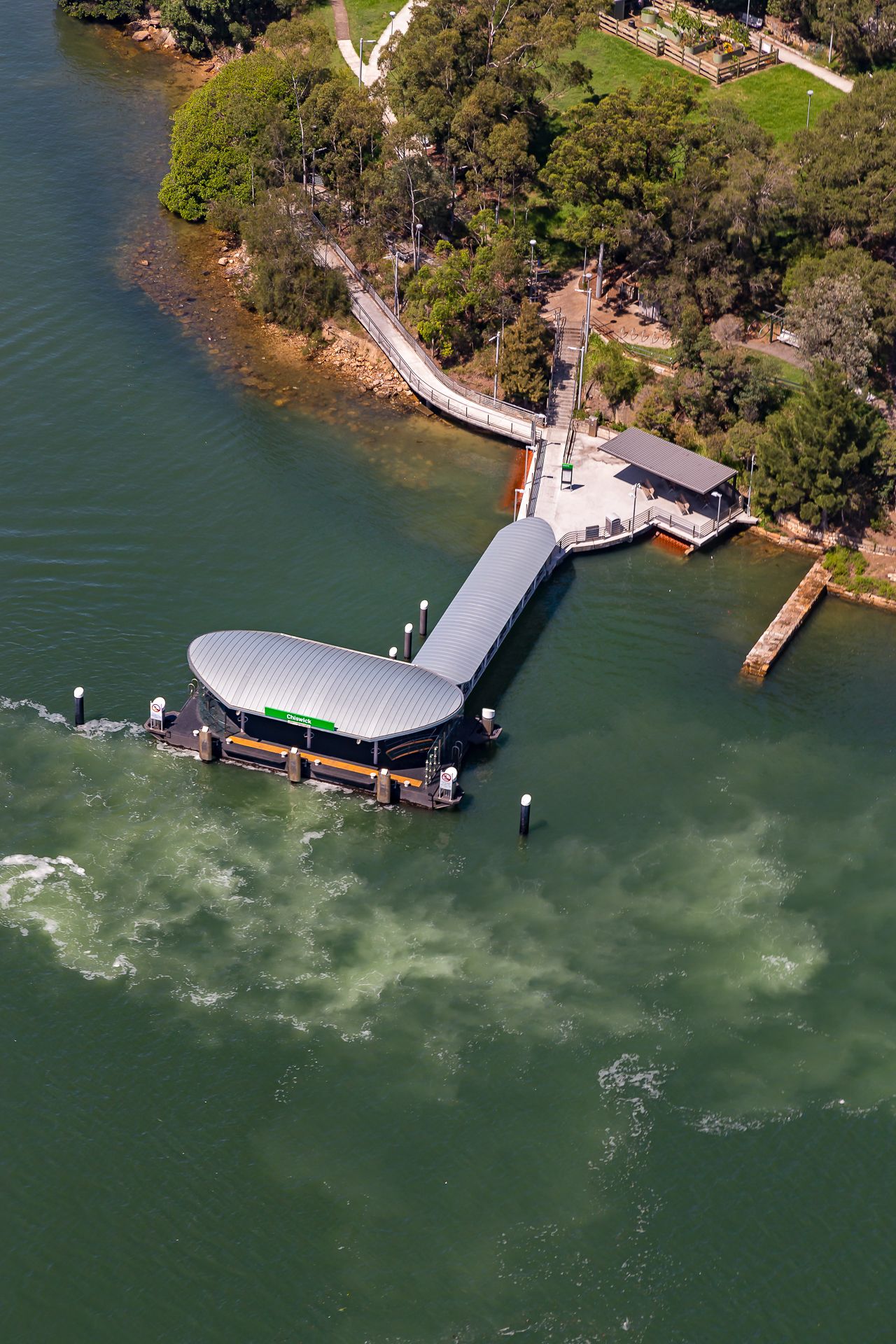 Aerial view of a curved-roof ferry wharf and walkway extending into green harbour water beside a leafy foreshore and winding pathway.