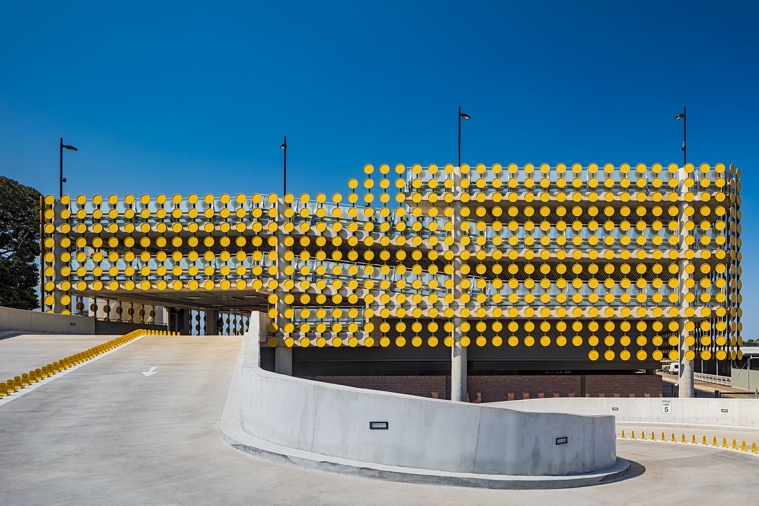 Modern parking structure with a bright yellow perforated facade under a deep blue sky, with a curved concrete ramp in the foreground.