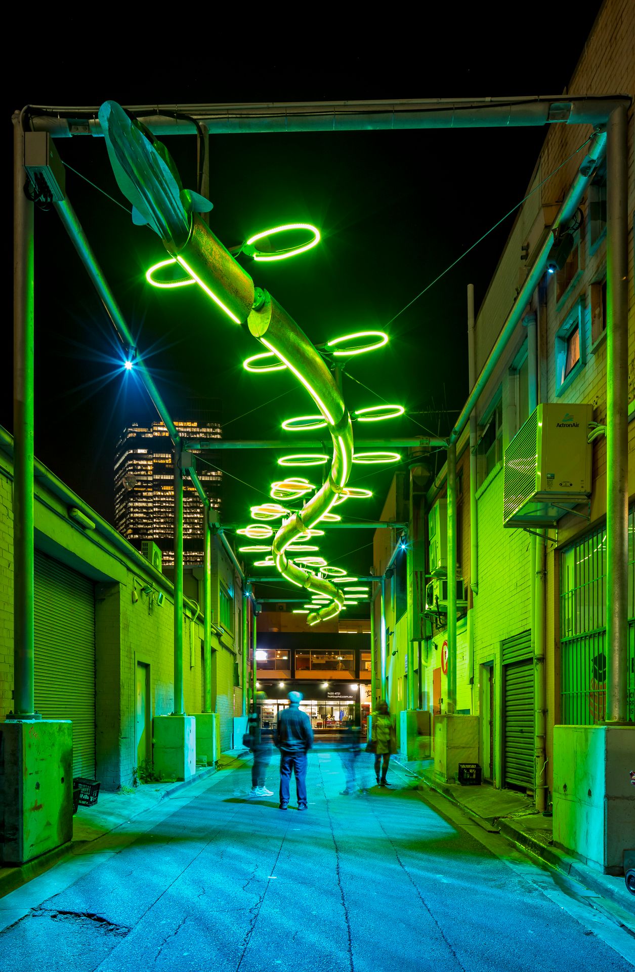 Green neon spiral light sculpture hangs above a narrow urban alley at night as pedestrians walk toward a glowing city skyline.