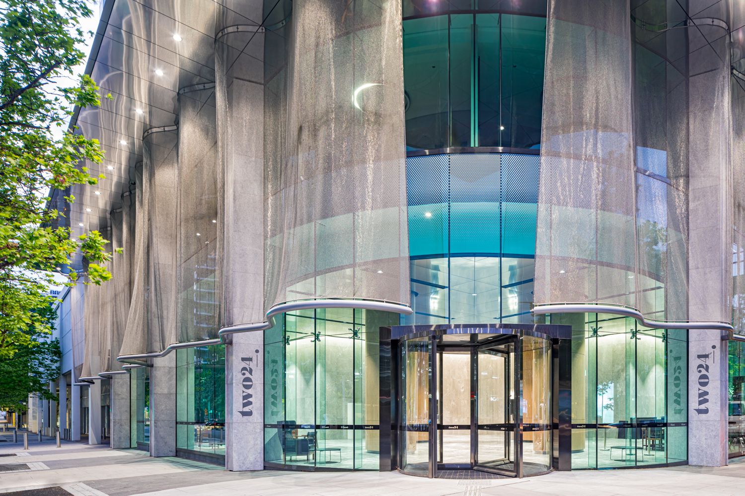 Modern glass and metal mesh office building entrance with revolving doors and trees in the foreground.
