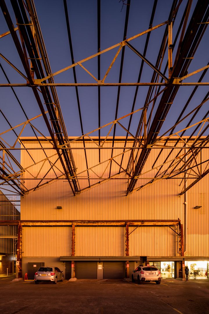 Large industrial warehouse with rusted steel roof trusses and a glowing orange corrugated facade against a deep blue twilight sky.
