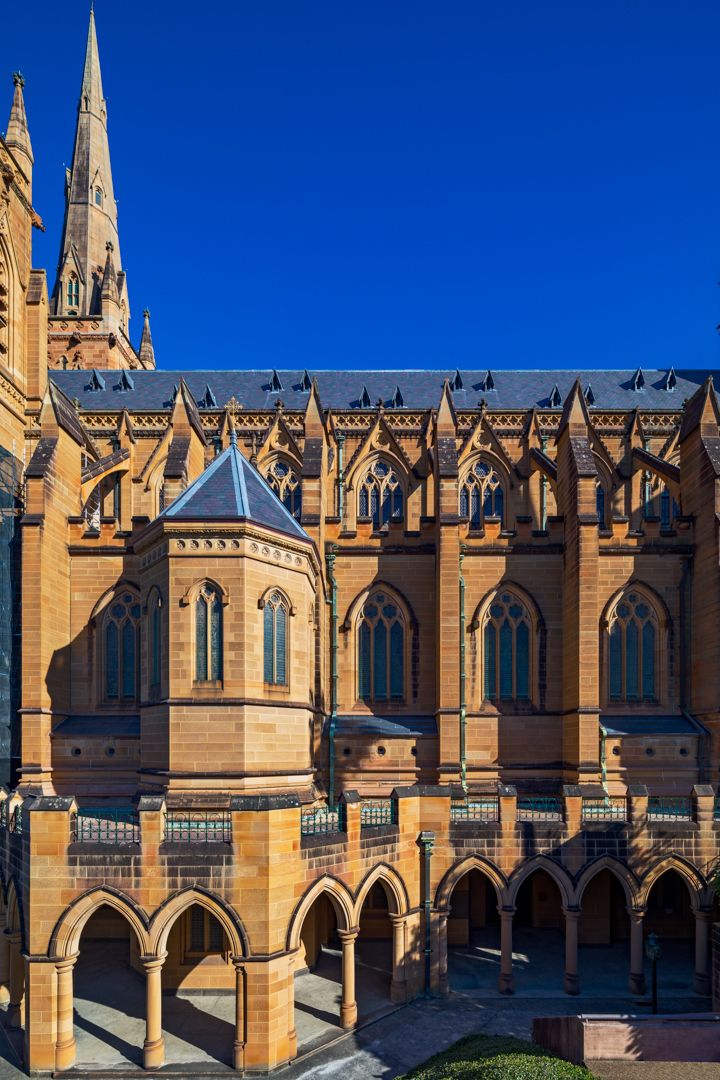 Gothic Revival sandstone cathedral facade with pointed arched windows, ornate spires and an arcaded cloister beneath a deep blue sky.