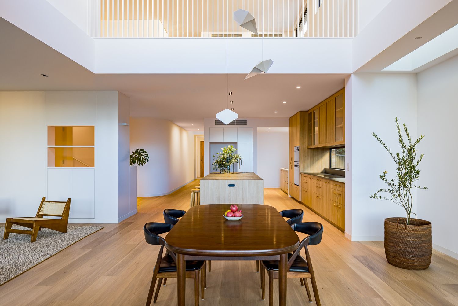 Bright double-height dining and kitchen space with a wooden table, black chairs, pendant lights and warm timber cabinetry.