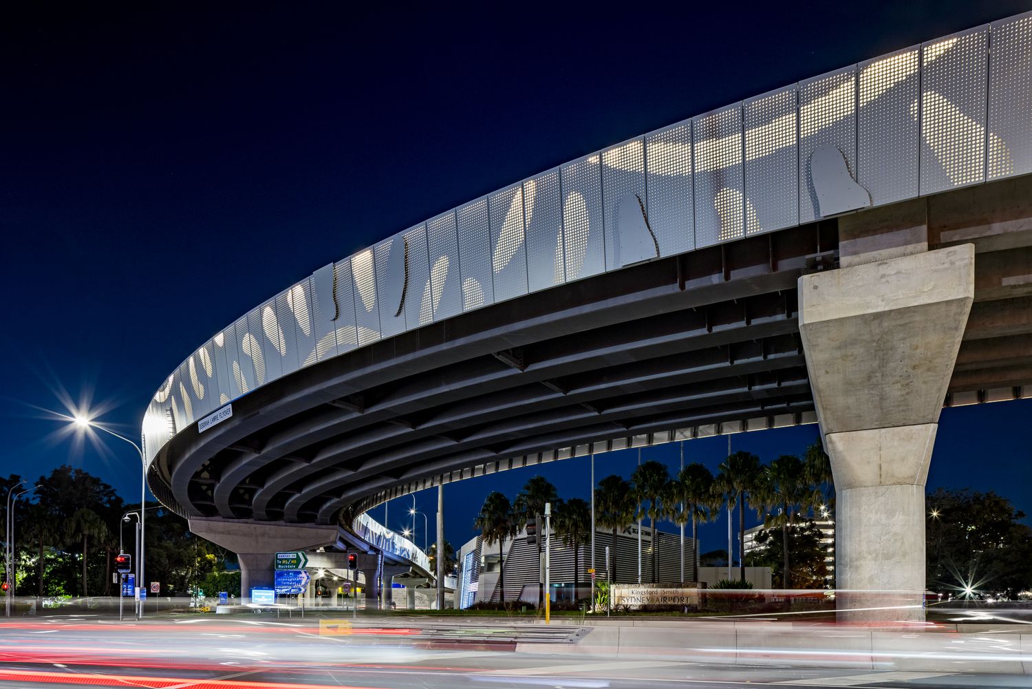 Underside of a curved elevated road with a patterned white facade, concrete pillars and palm trees at dusk.