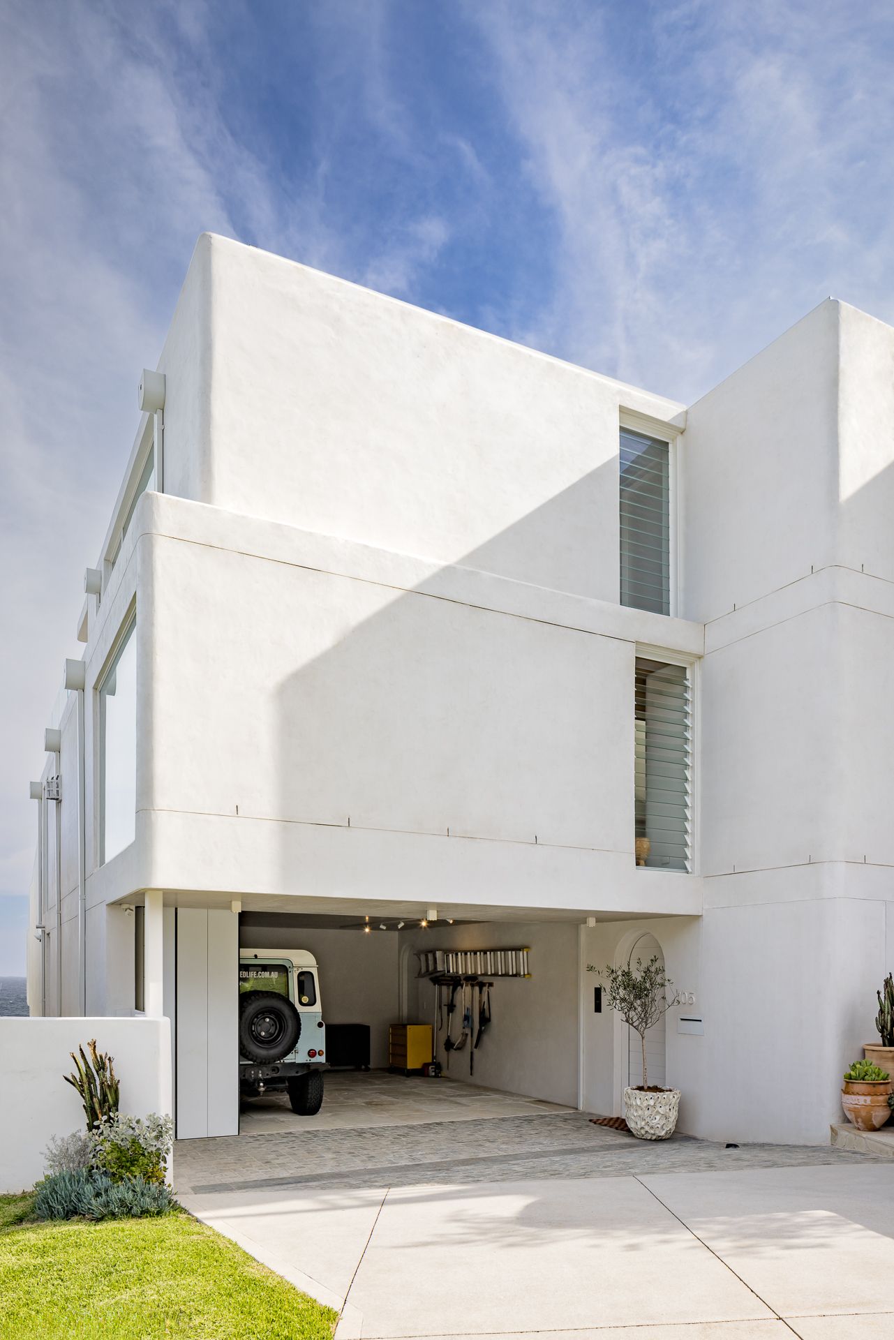 White contemporary beach house with open garage showing a vintage 4x4 vehicle, louvered windows and a landscaped driveway.