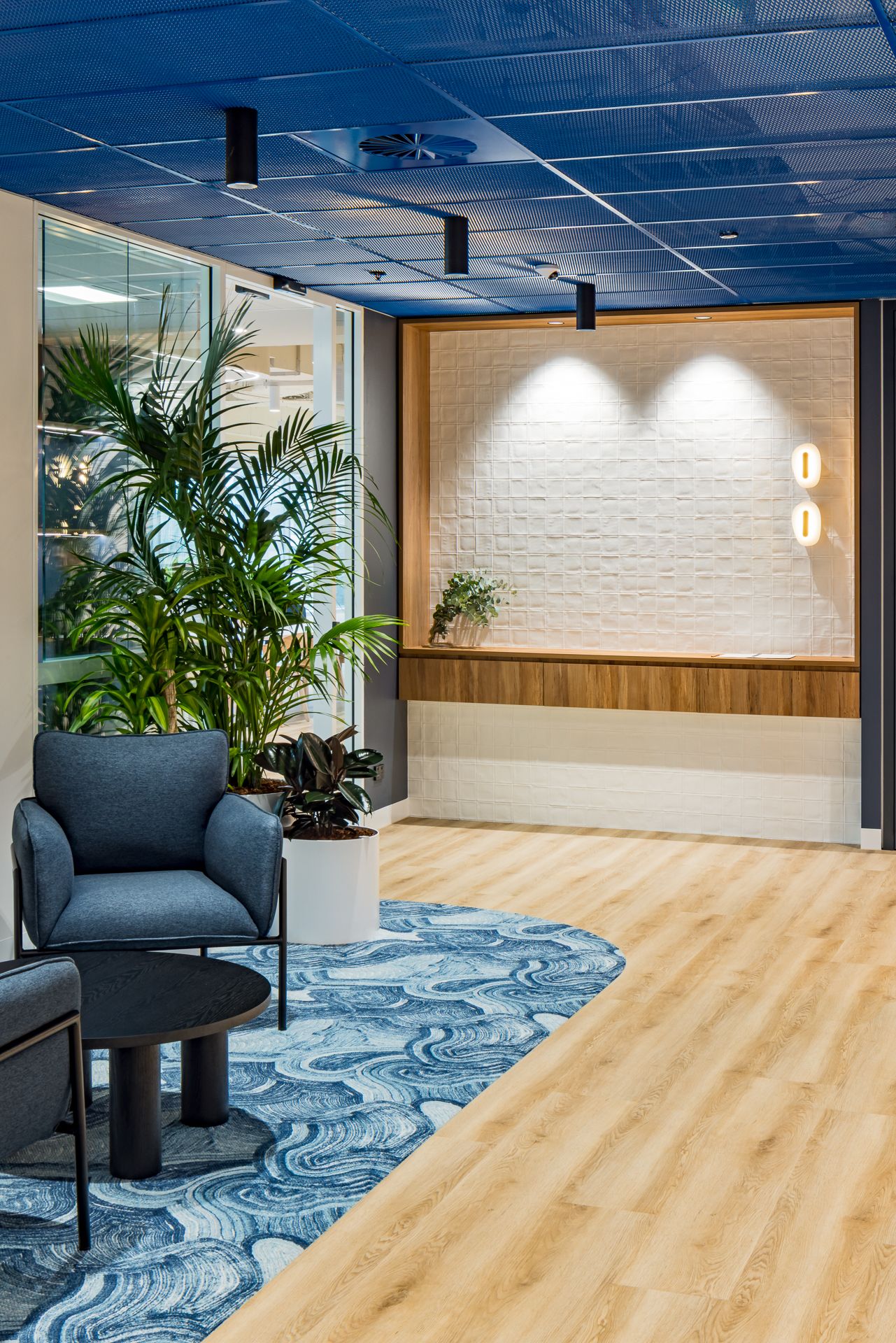 Modern office lounge with blue armchair, round black table, potted palms, wood flooring and a textured white feature wall.