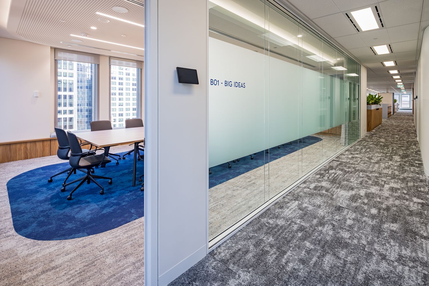 Office corridor with patterned carpet and frosted glass walls leading past a meeting room with a round table and blue rug.