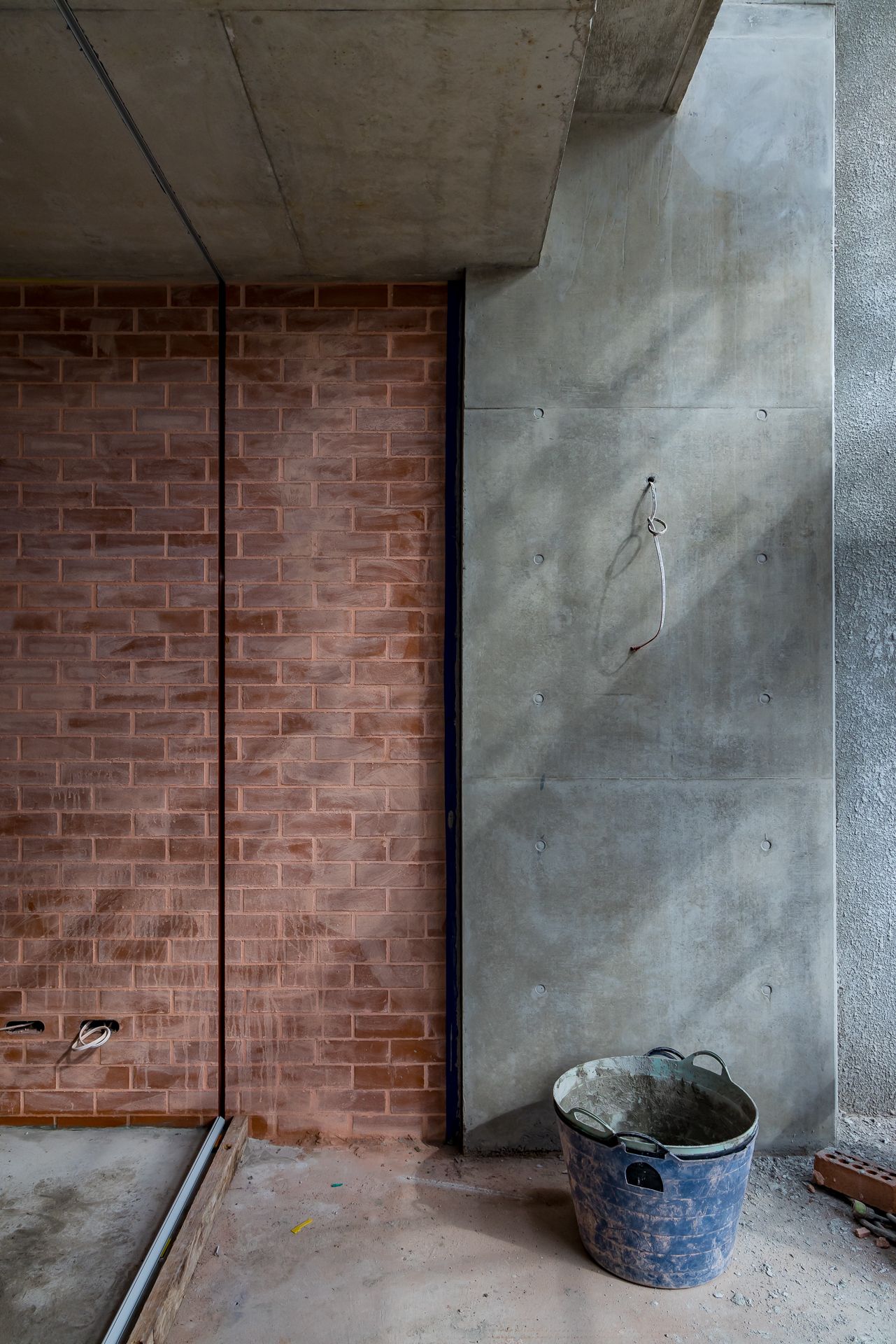 Raw concrete wall meeting a red brick wall in a construction site, with a blue plastic bucket on the dusty floor.