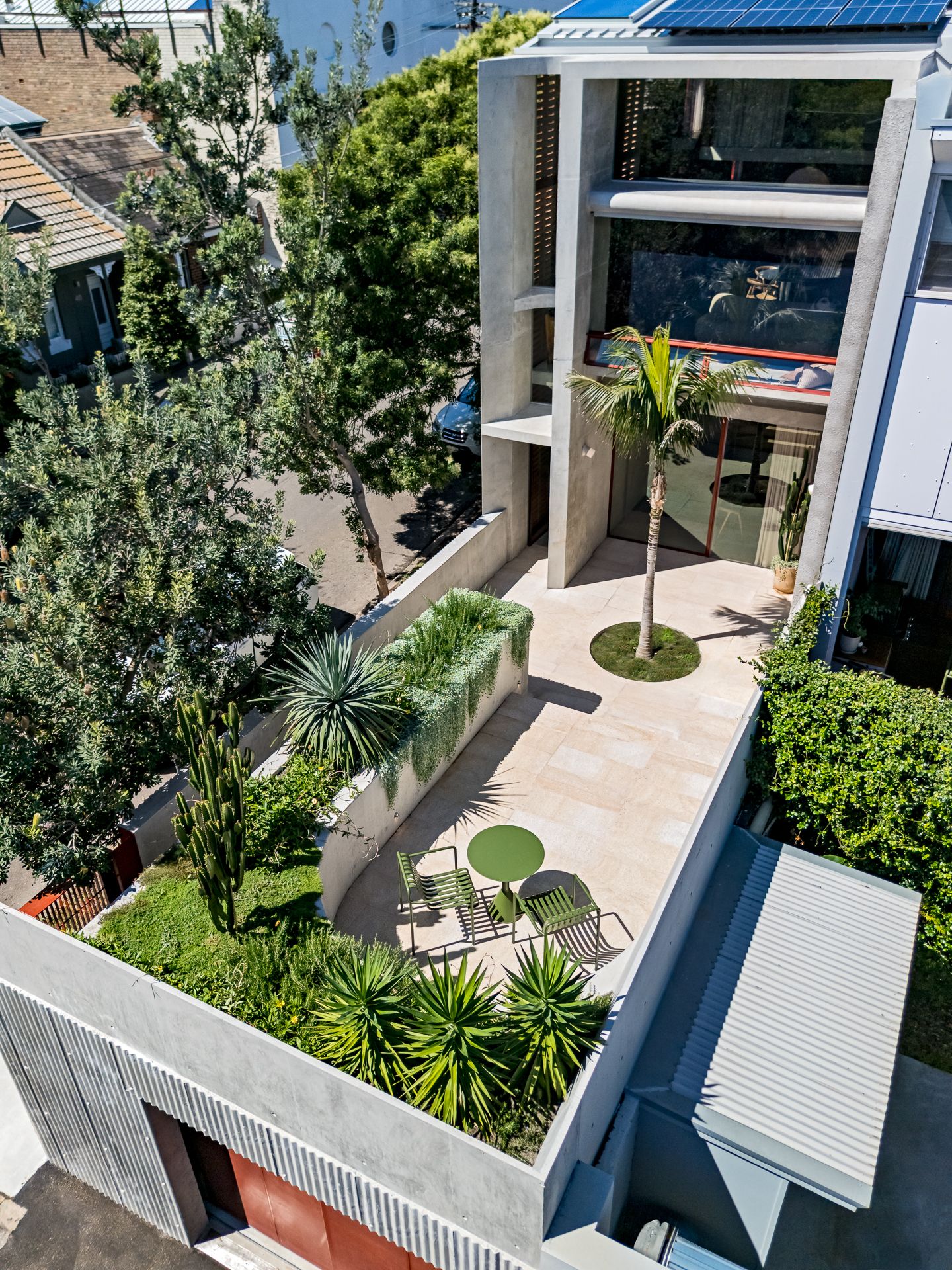 Elevated view of a modern concrete home with a palm courtyard, garden beds, a green table and chairs, and rooftop solar panels.