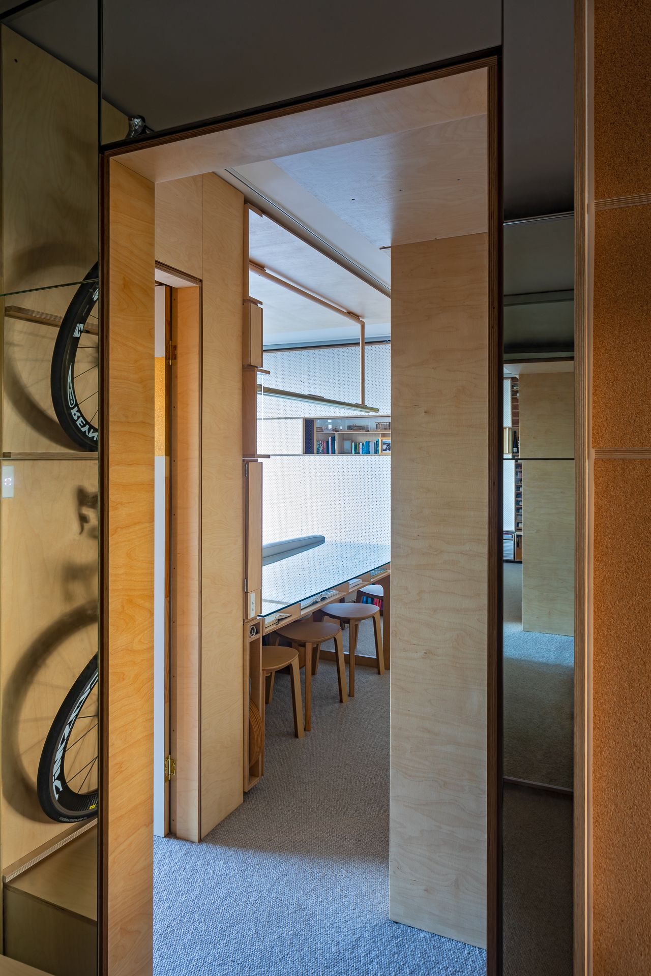 View through a plywood-framed doorway into a bright workspace with a long desk and wooden stools, with bicycle wheels reflected in mirrors.