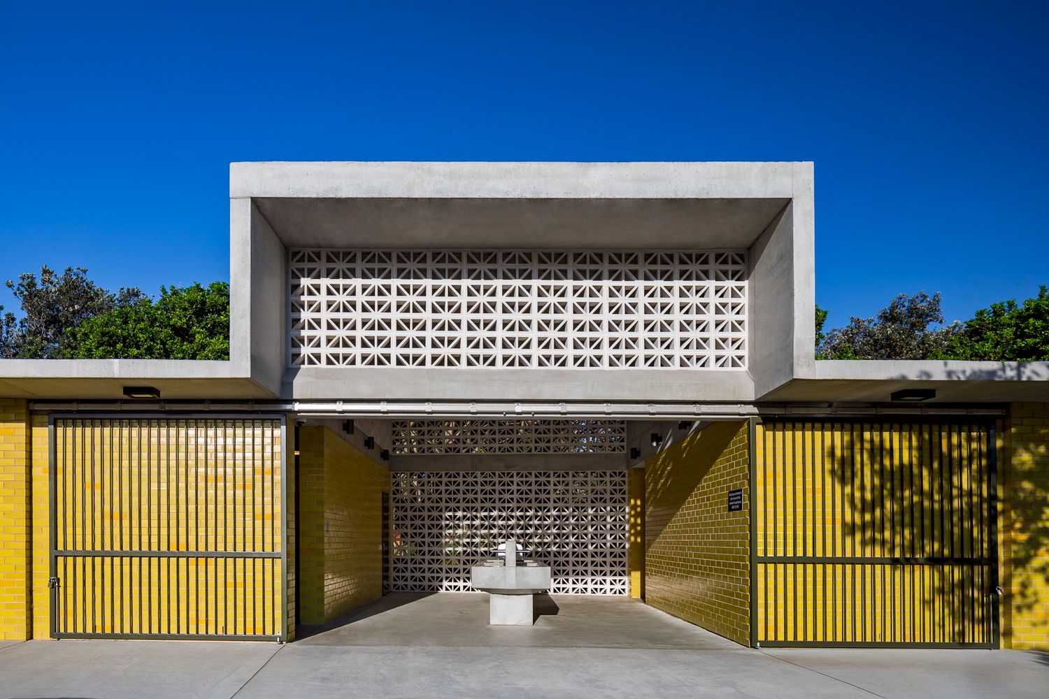 Modern single-story building with a concrete facade, decorative breeze block screen, and bright yellow sliding gates under a clear blue sky.