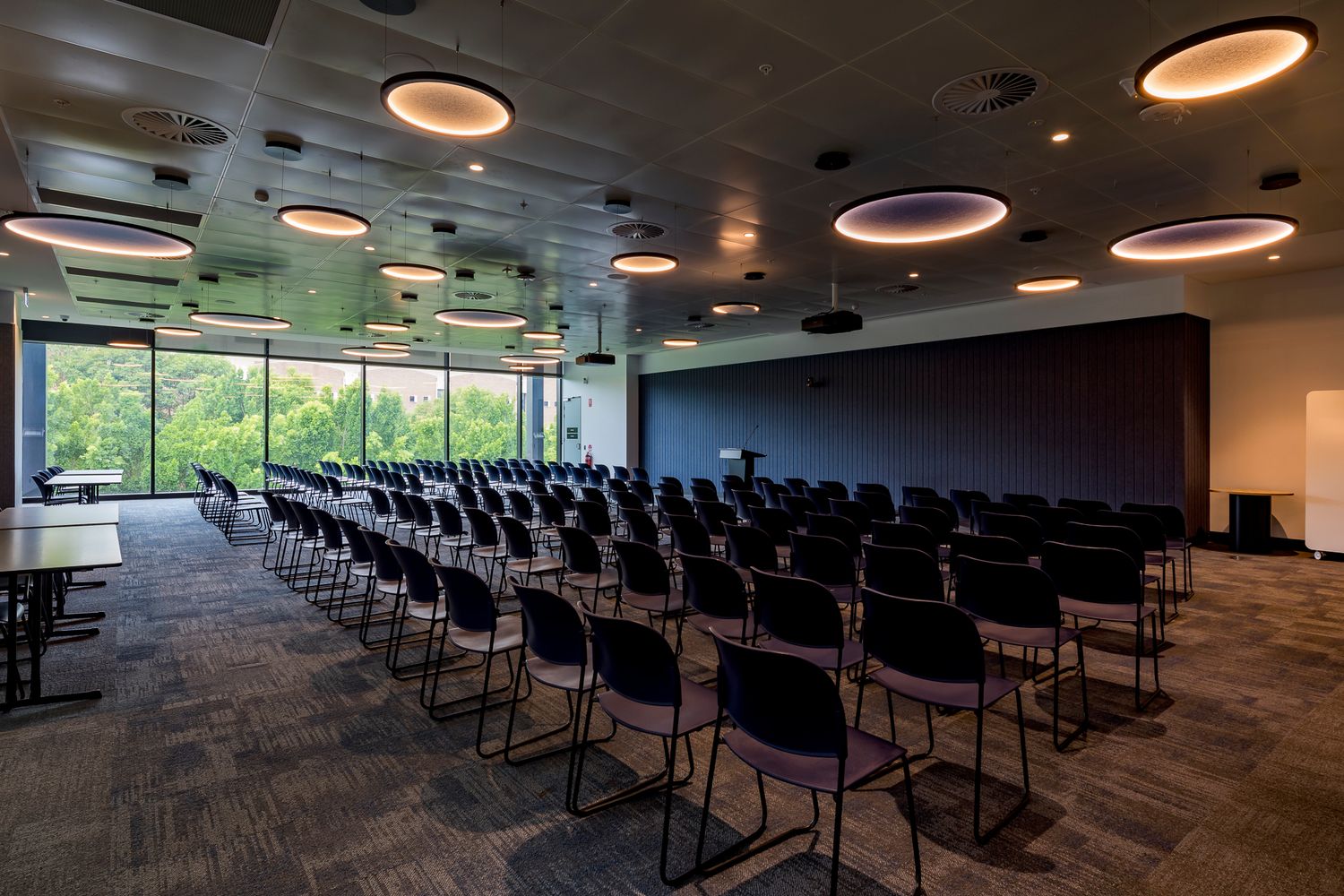 Large lecture room with rows of black chairs, circular ceiling lights and a wall of windows overlooking trees.
