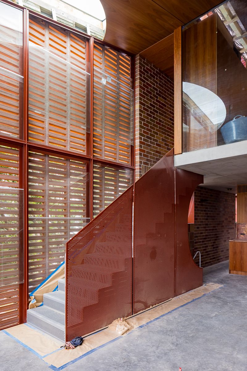 Interior staircase with perforated rust-red metal balustrade, timber louvered windows, exposed brick walls and a concrete mezzanine.