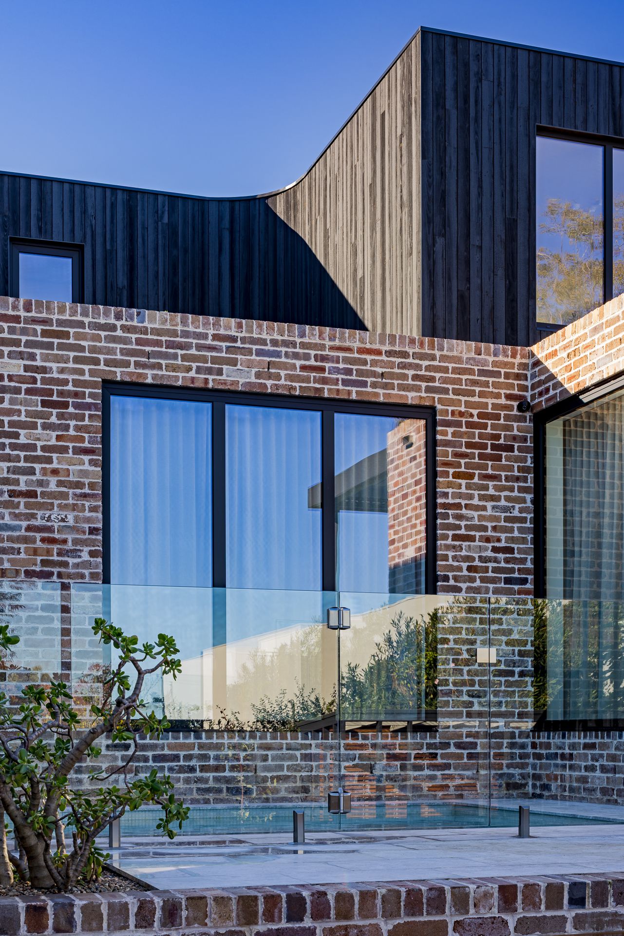 Detail of a modern home combining recycled red brick, dark timber cladding, black-framed windows and a glass pool fence.