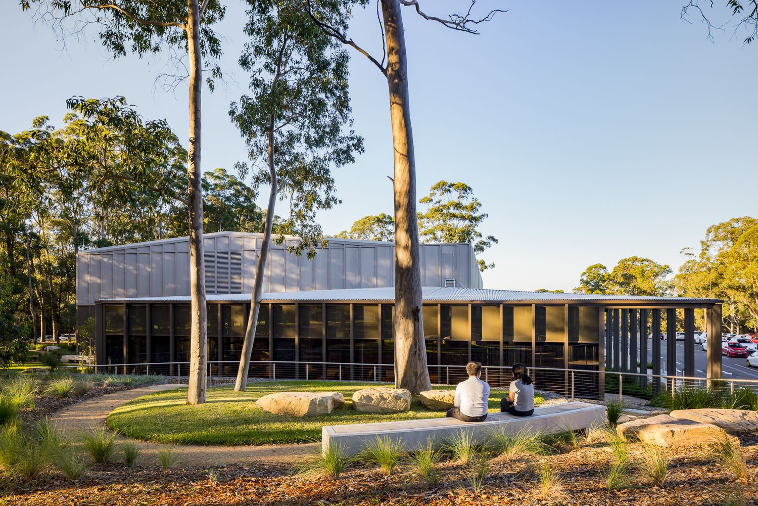 Two people sit on a low concrete bench in a landscaped garden in front of a modern glass and steel building among eucalyptus trees.