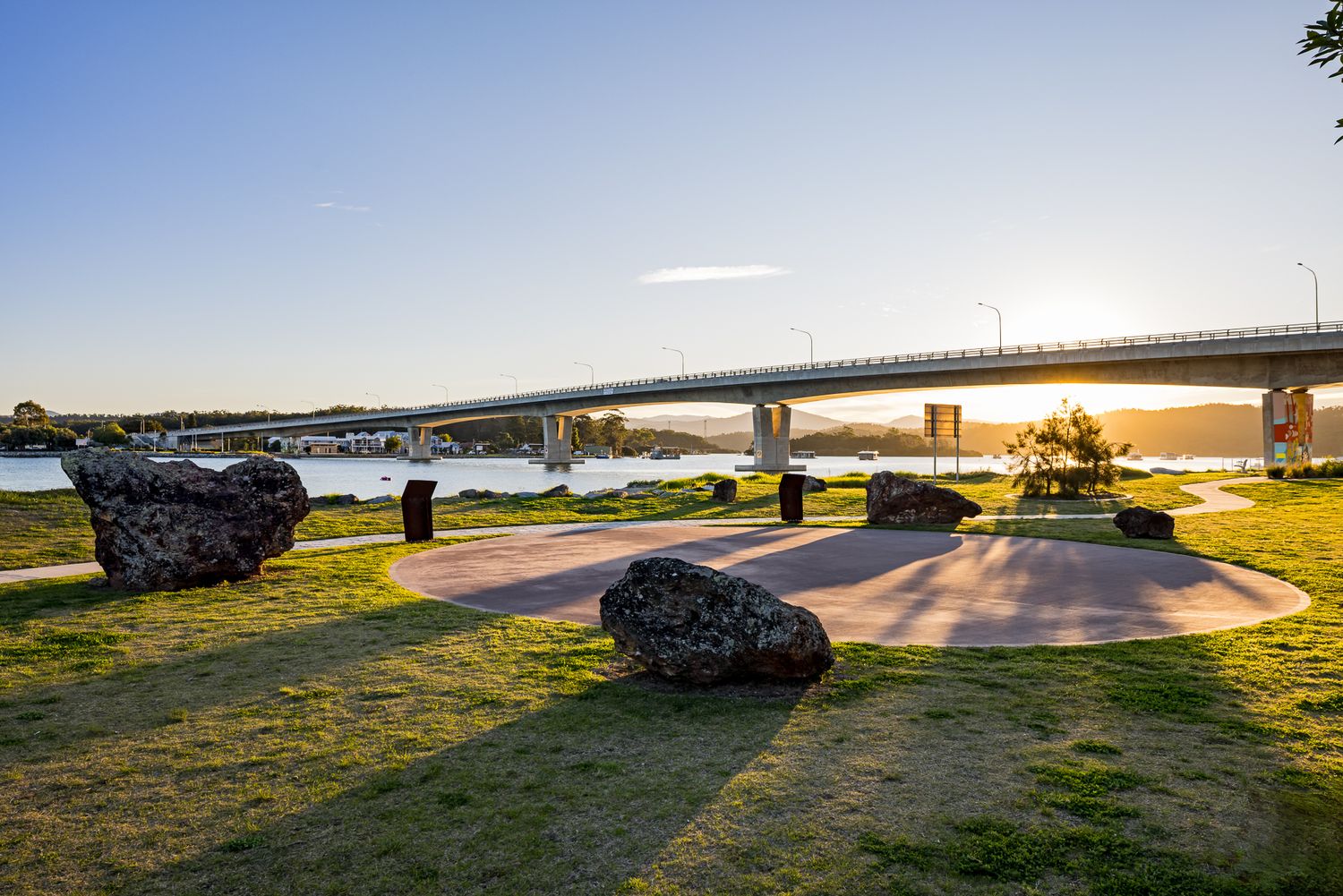 Sunlit waterfront park with large boulders and a circular paved area, with a long road bridge crossing the bay in the distance.