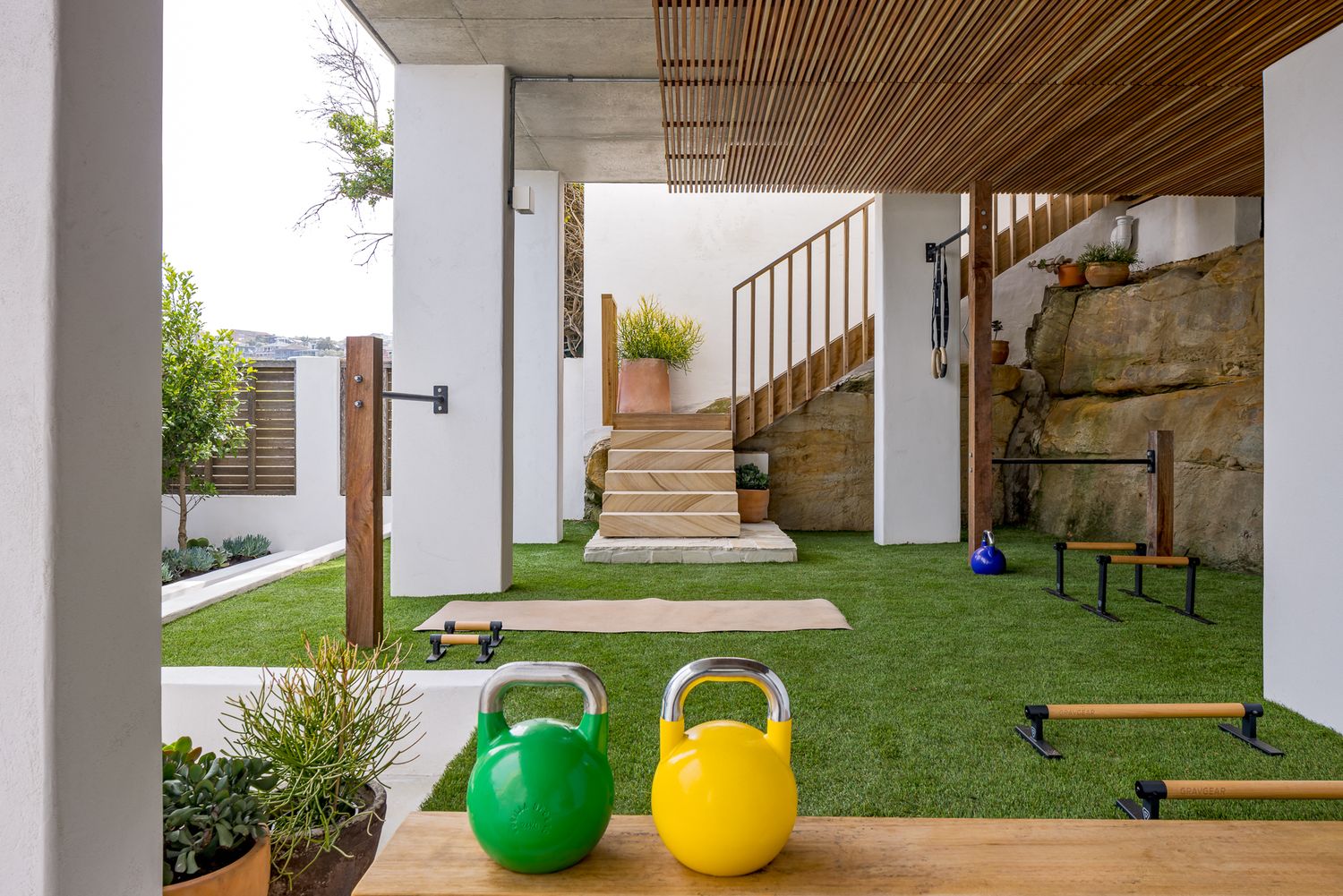 Landscaped garden gym with turf, green and yellow kettlebells, timber pergola, stone steps and outdoor training equipment.