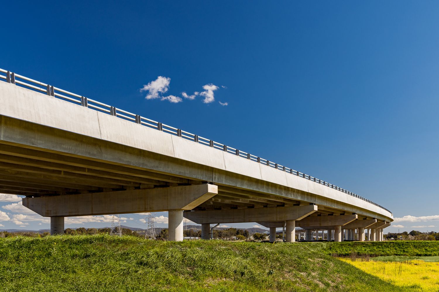 Elevated concrete highway bridge curving over green grassland and yellow flowers beneath a vivid blue sky with small white clouds.