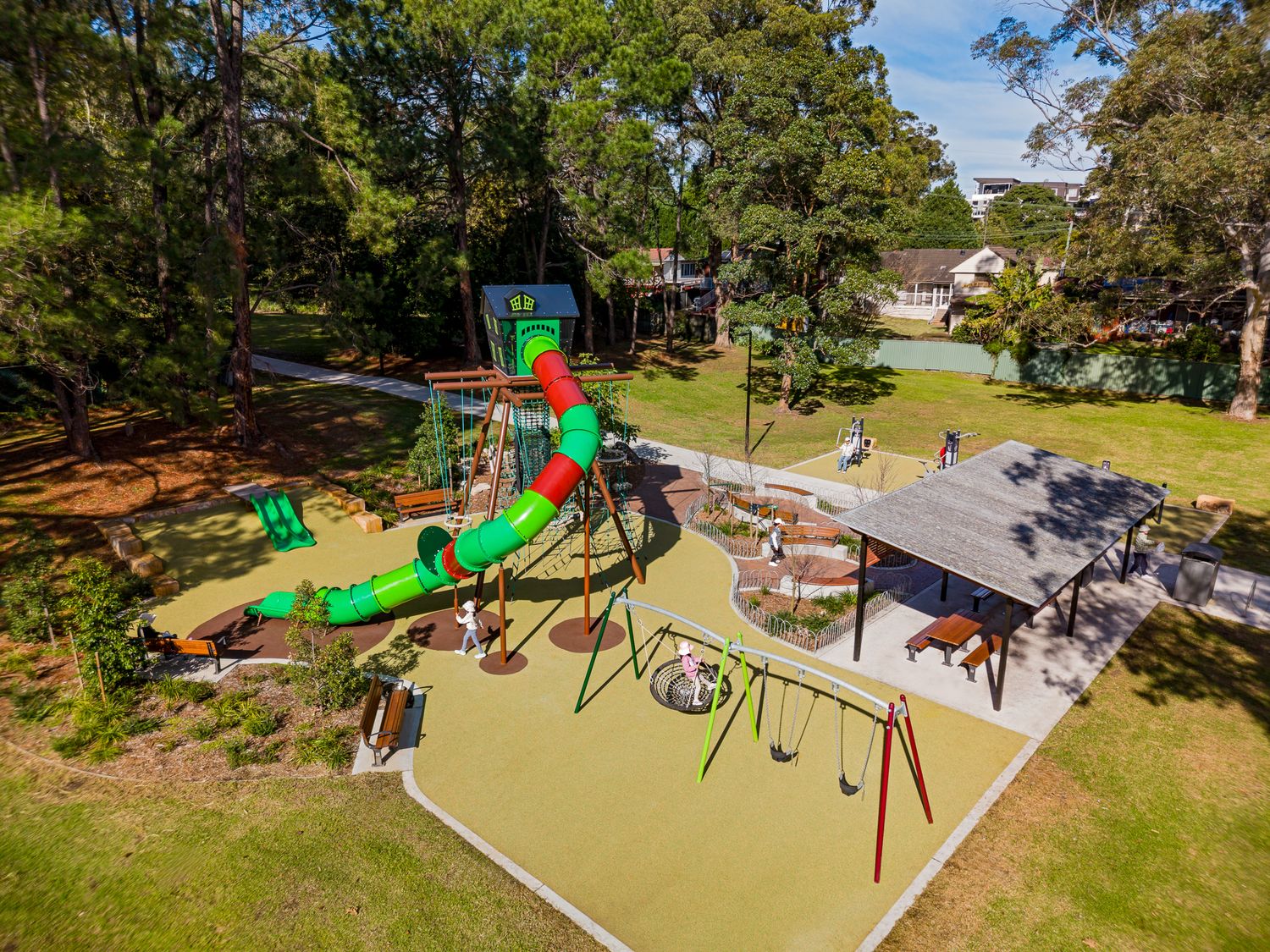 Aerial view of a colourful park playground with a spiral tube slide, swings, shaded picnic shelter and seating among tall trees and grass.