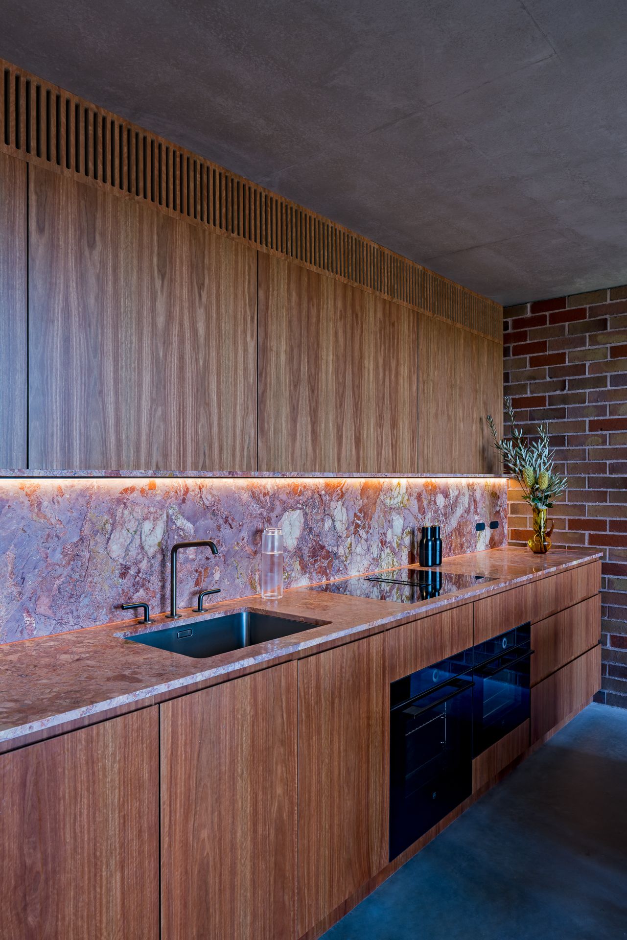 Galley kitchen with timber cabinets, pink marble splashback and benchtop, black tapware and a concrete ceiling beside brick.