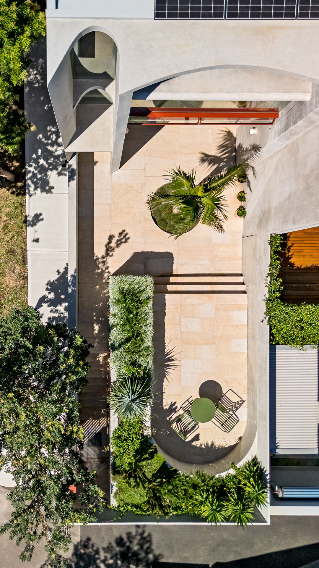 Aerial view of a courtyard with a palm tree, sandstone paving, a green garden bed, and a round table with chairs beside solar panels.