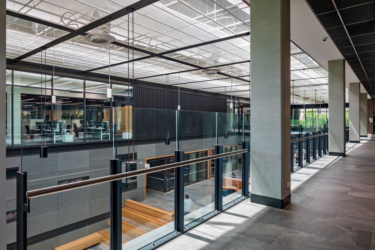 Spacious campus interior walkway with glass balustrade, concrete columns and a view over an open-plan study area below.